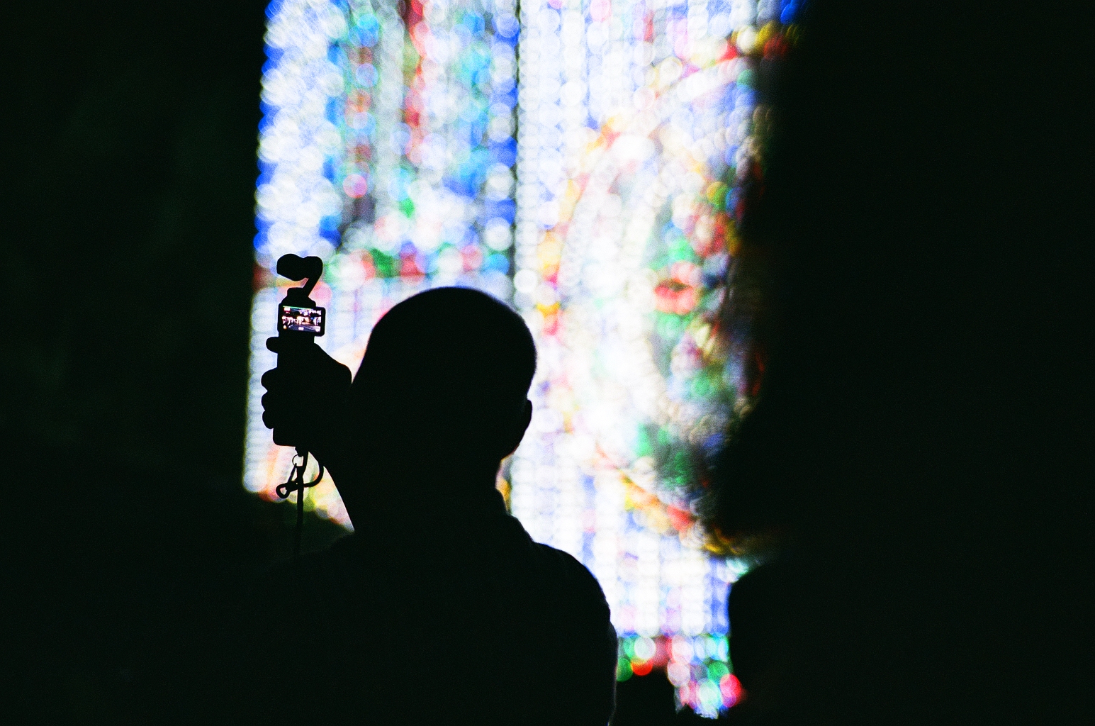 Photo of a man holding up a small camera to take a video of his surroundings, silhouetted against the large light installation