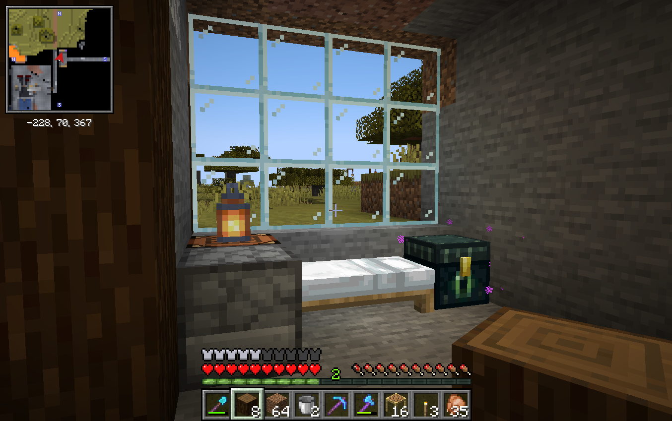 A screenshot showing a small, cozy room. There is a large glass window with a bed in front of it. Next to the bed are an ender chest, a crafting table with a lantern on it, and a furnace.