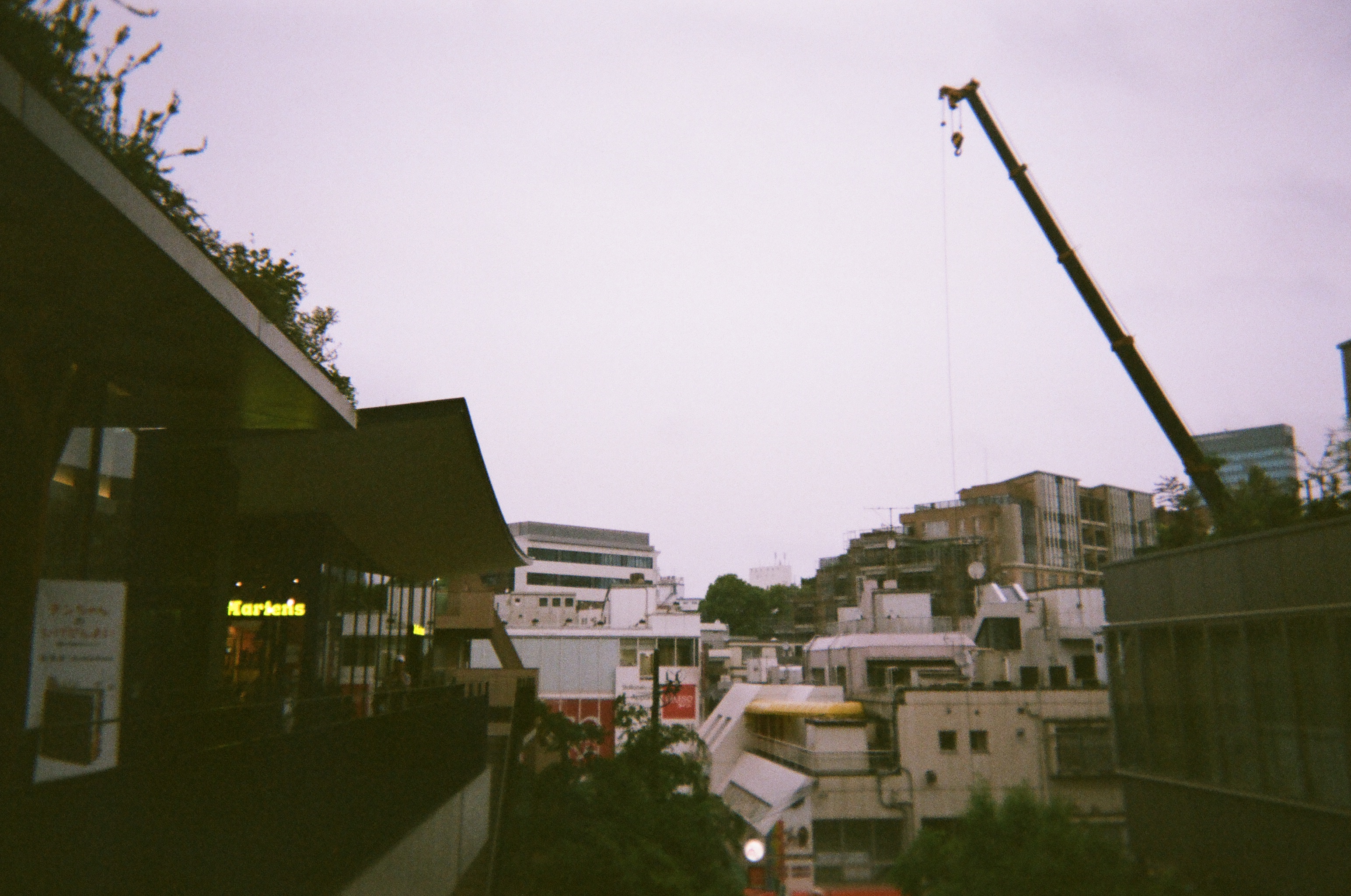 Photo from above ground level on a rainy day. The sky is a milky greyish-white. There are many shorter buildings below, but a single long construction crane extends into the sky.