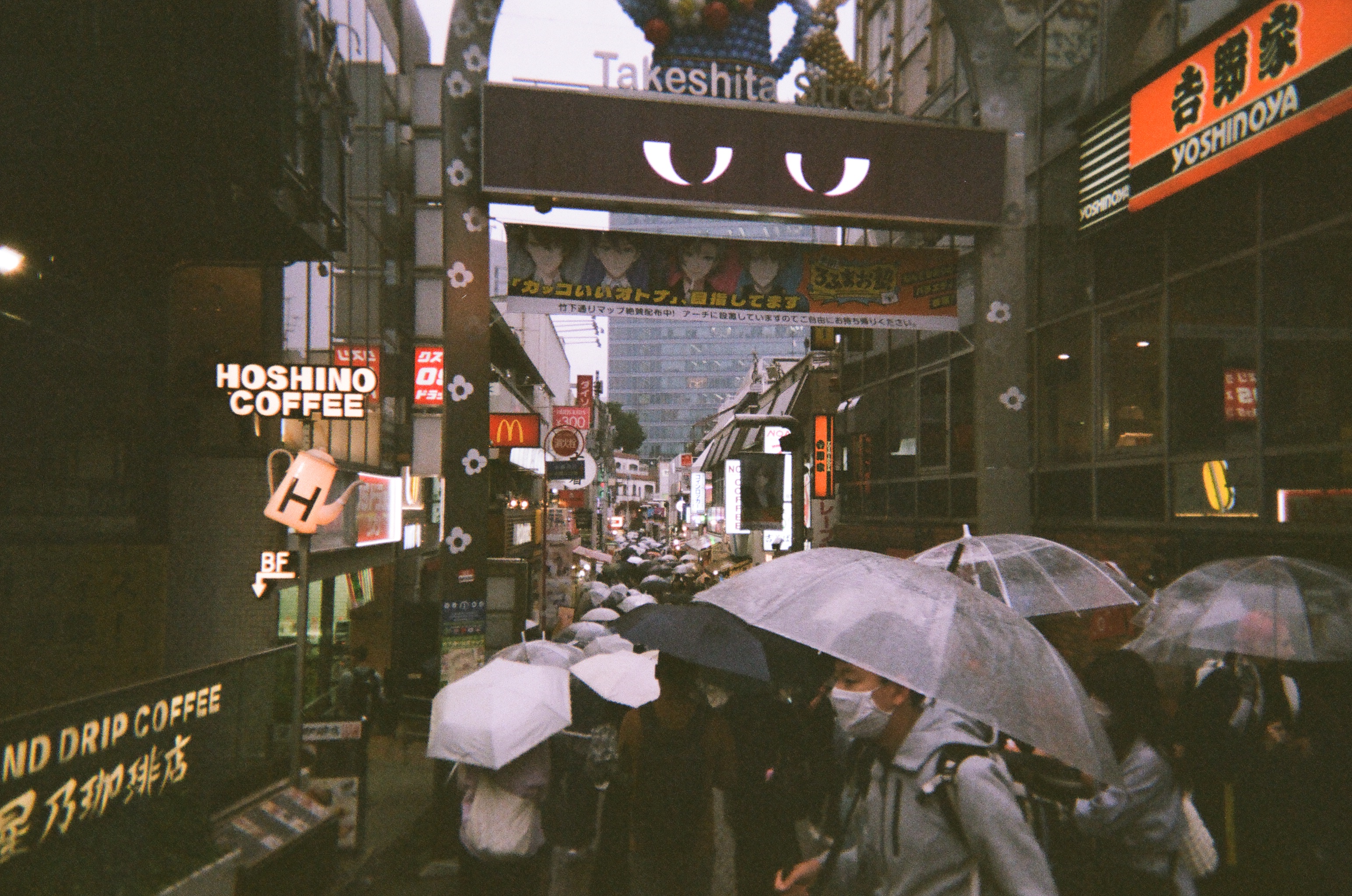 Photo of a narrow pedestrian street flanked by various signs and shops. The street is packed with people carrying umbrellas. A signboard above reads "Takeshita Street" with a digital screen showing a pair of half-lidded unimpressed eyes underneath.