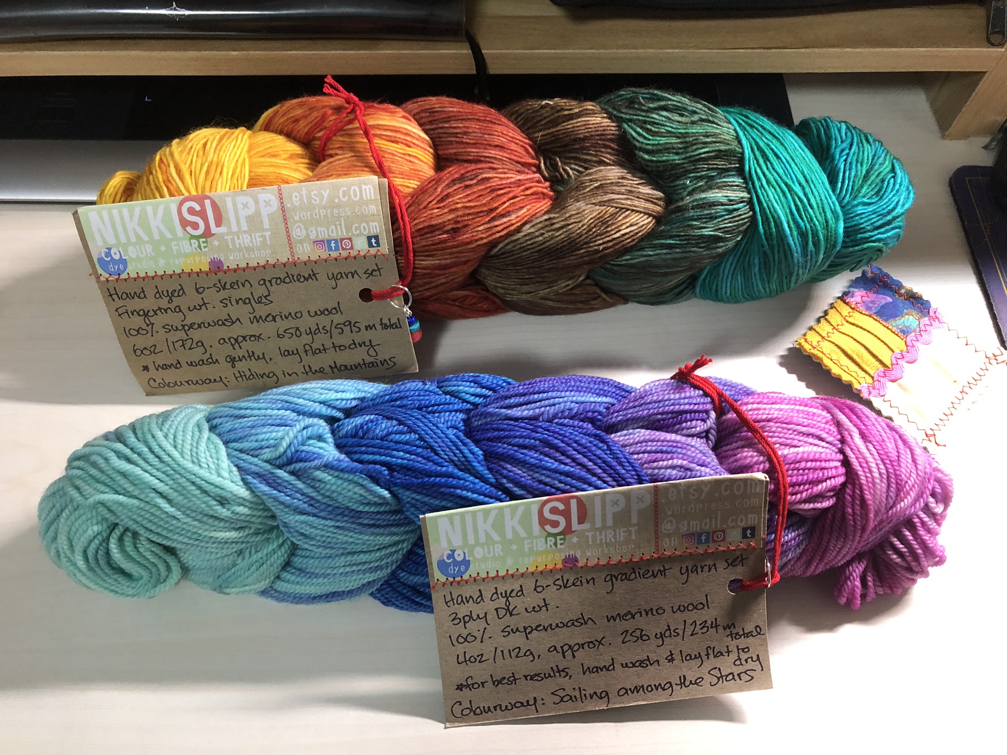 A photo of 2 bundles of yarn that look like large braids. Each bundle has 6 colours of yarn in small skeins, arranged in a pretty gradient. The top gradient goes from yellow to red to brown to teal, and the bottom gradient goes from light blue to darker blue to purple to pink. Both have the dyer's labels attached in cardboard.