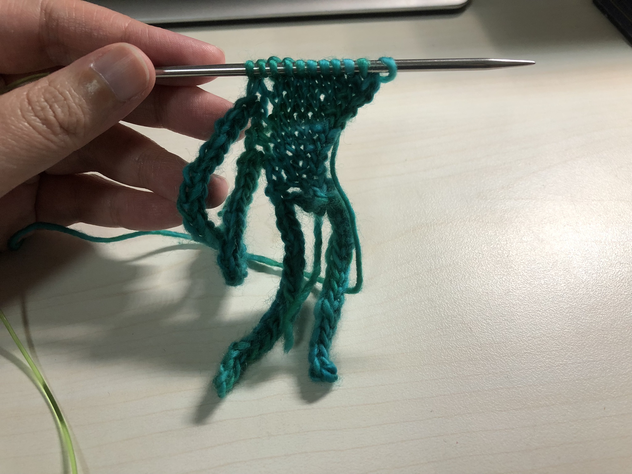 Photo of a knitting project on a knitting needle, held horizontally in a hand. It looks like a small triangle with four long tassels coming off the sides.