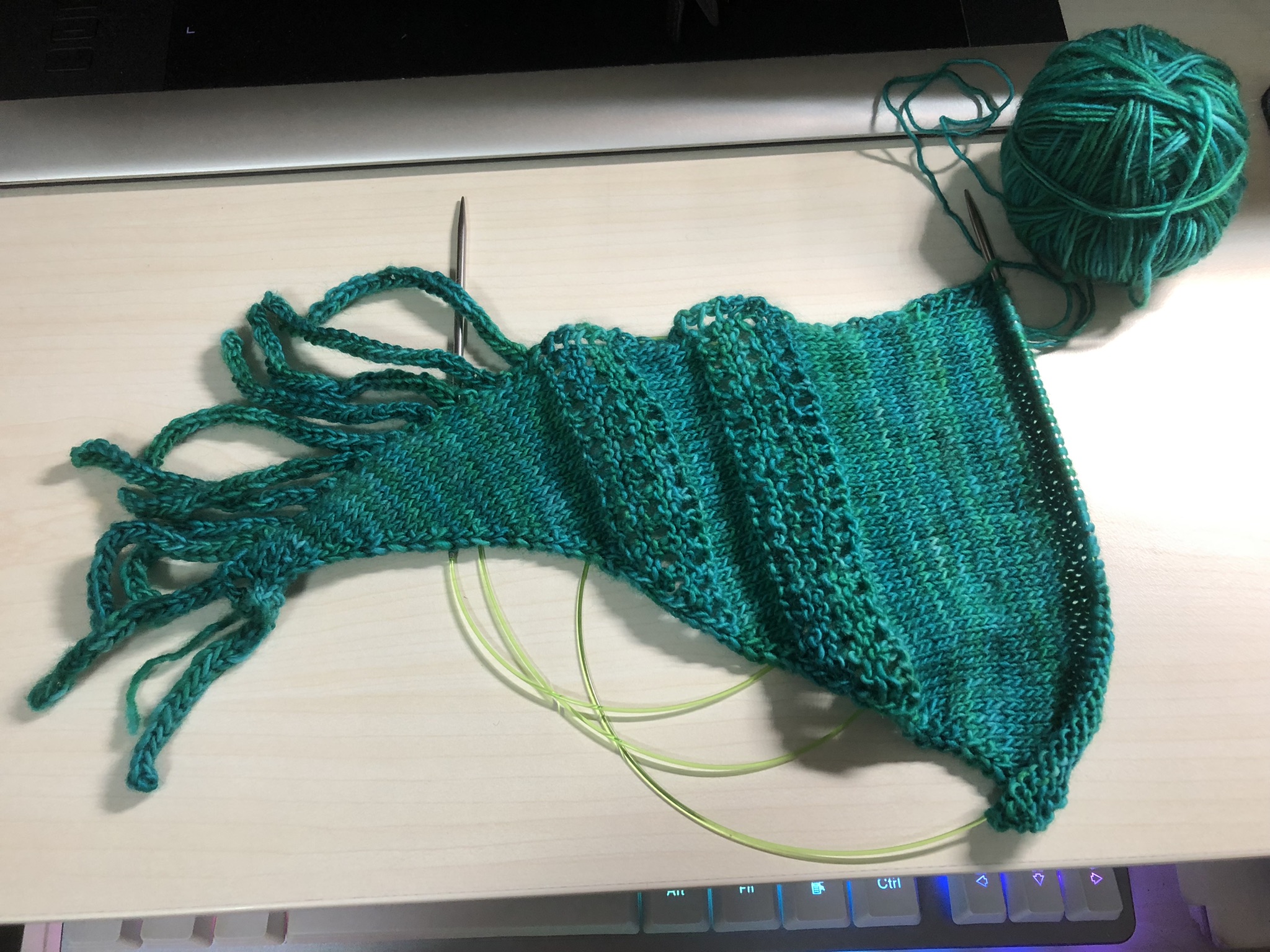 Photo of a knitting project on the needles. It is triangular in shape, knitted in teal yarn with some tassels at the end.