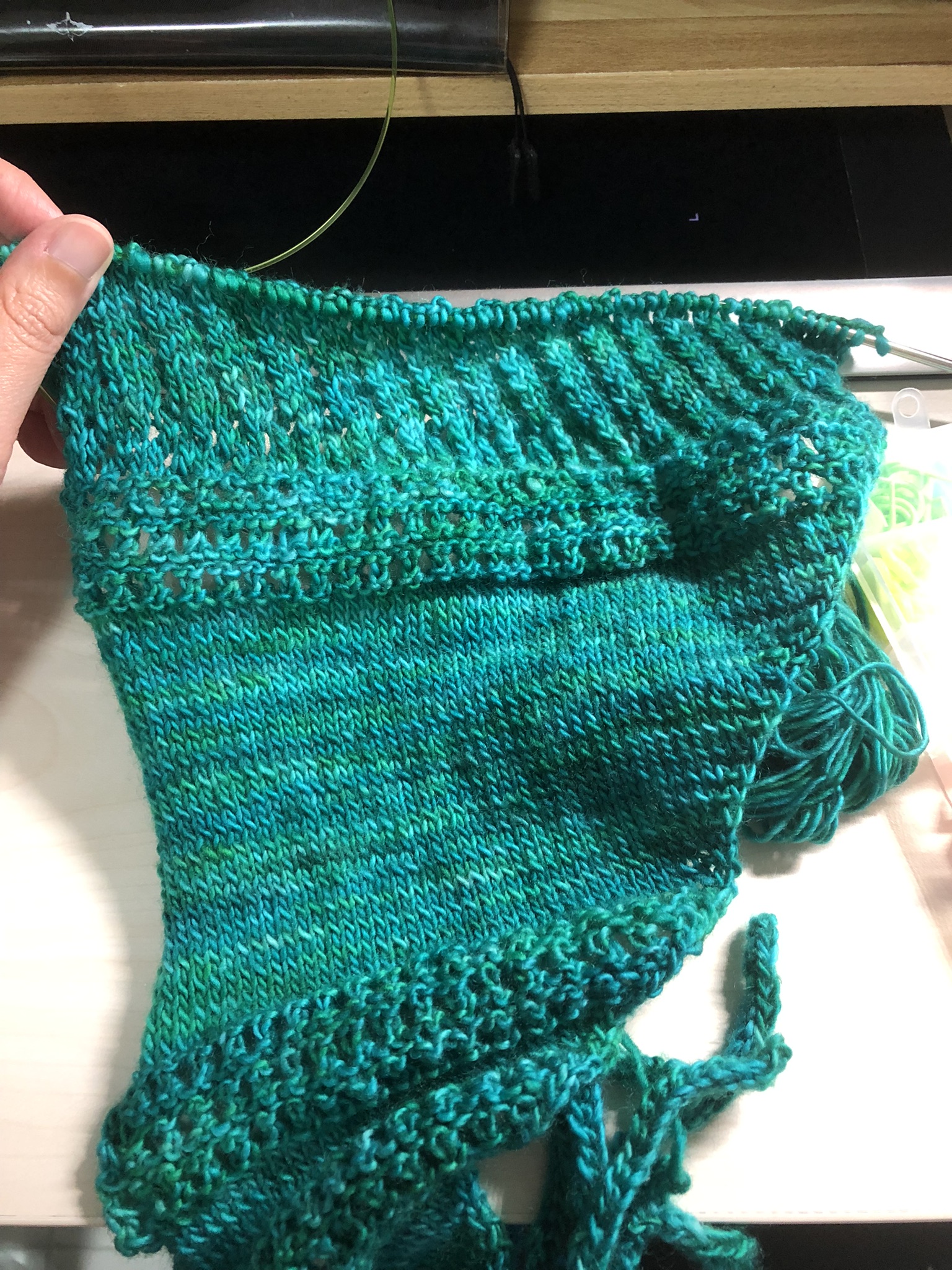 Photo of a knitting project on the needles. It is triangular in shape, knitted in teal yarn with some tassels at the end.