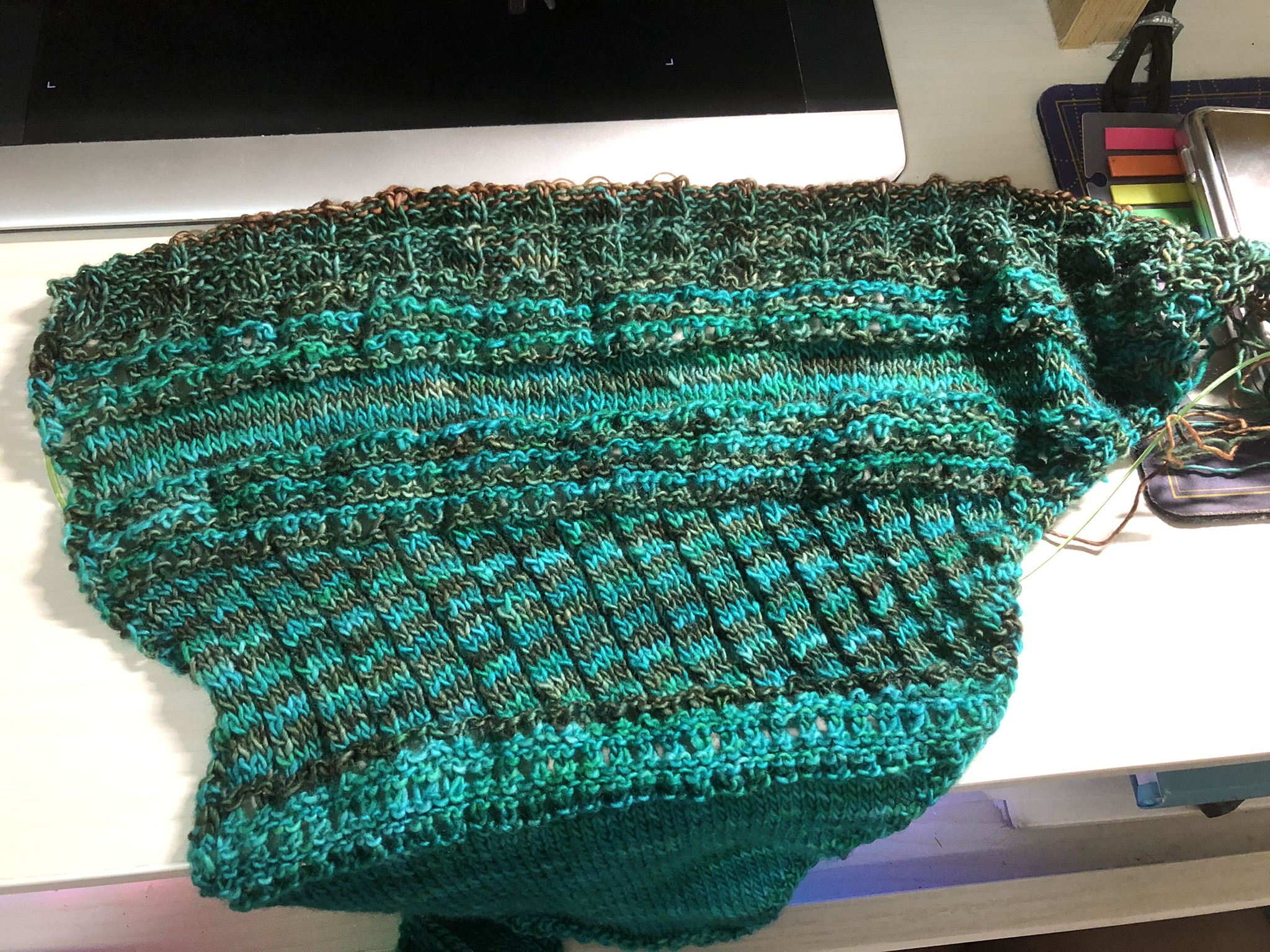 Photo of a knitting project on the needles. It is triangular in shape, knitted in teal yarn with some tassels at the end. Greenish brown yarn has been added in stripes in the newest sections.