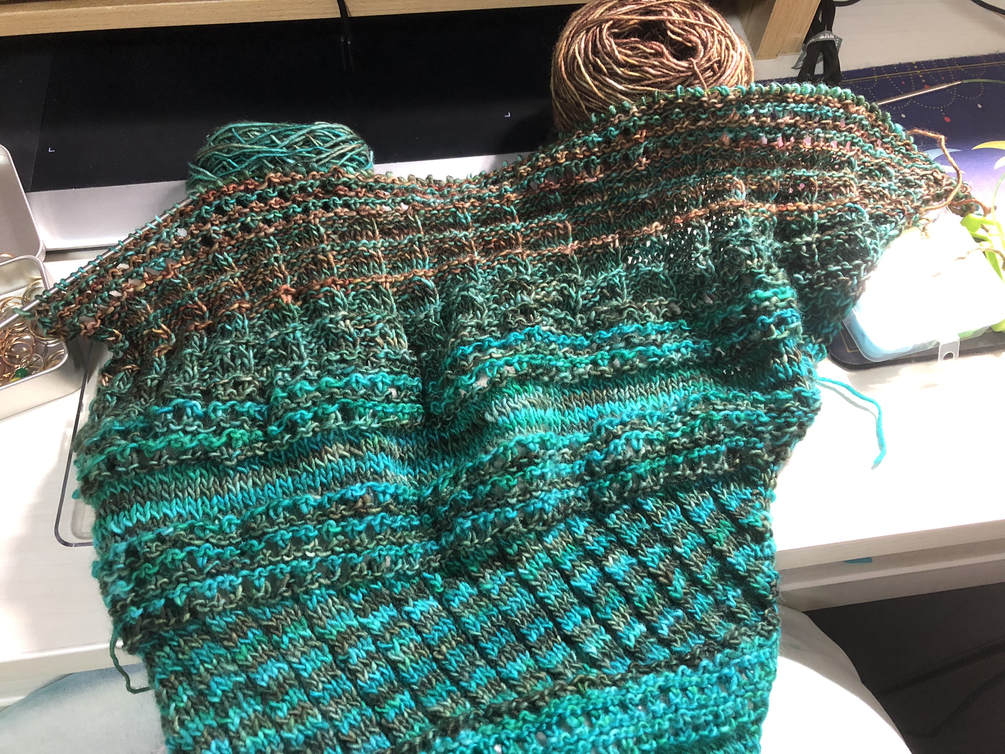 Photo of a knitting project on the needles. It is triangular in shape, knitted in stripes in teal and greenish brown yarn. Reddish-brown yarn has been added in stripes in the newest sections.