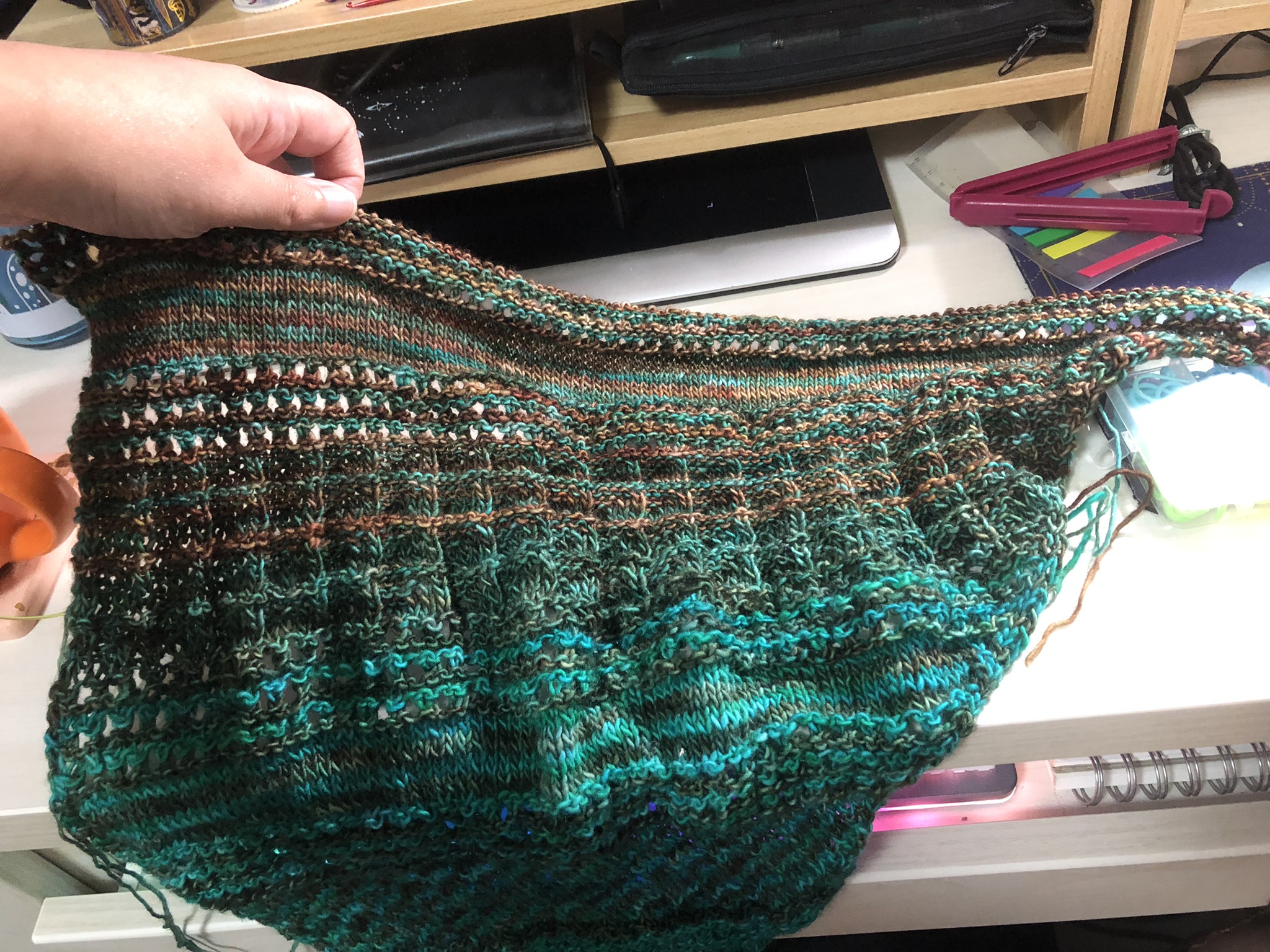 Photo of a knitting project on the needles. It is triangular in shape, knitted in stripes in teal and greenish brown yarn. Reddish-brown yarn has been added in stripes in the newest sections.