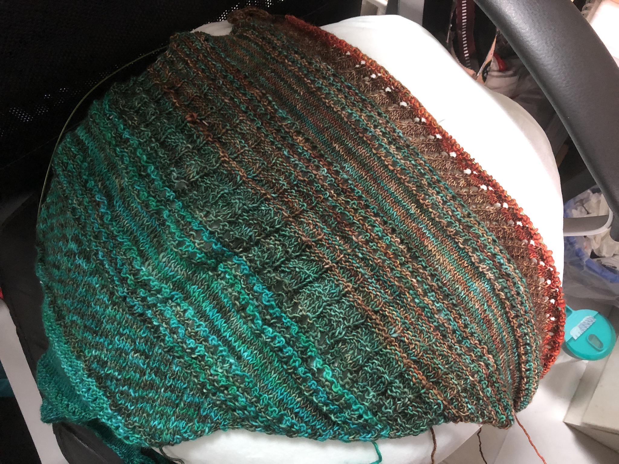 Photo of a knitting project on the needles. It is triangular in shape, knitted in stripes in teal, greenish brown, and reddish-brown yarn. Reddish yarn has been added in stripes in the newest sections.