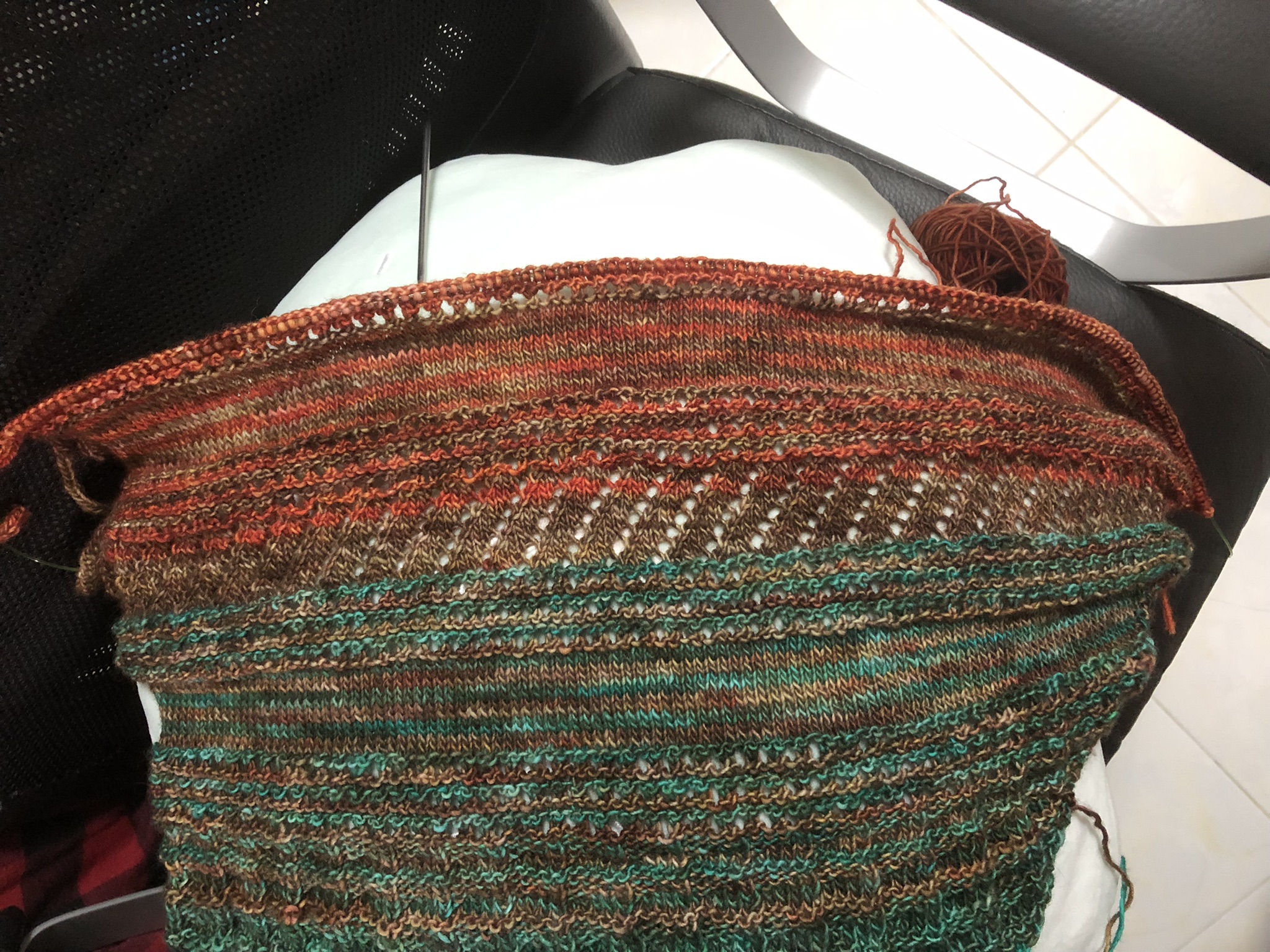 Photo of a knitting project on the needles. It is triangular in shape, knitted in stripes in teal, greenish brown, and reddish-brown yarn. Reddish yarn has been added in stripes in the newest sections.