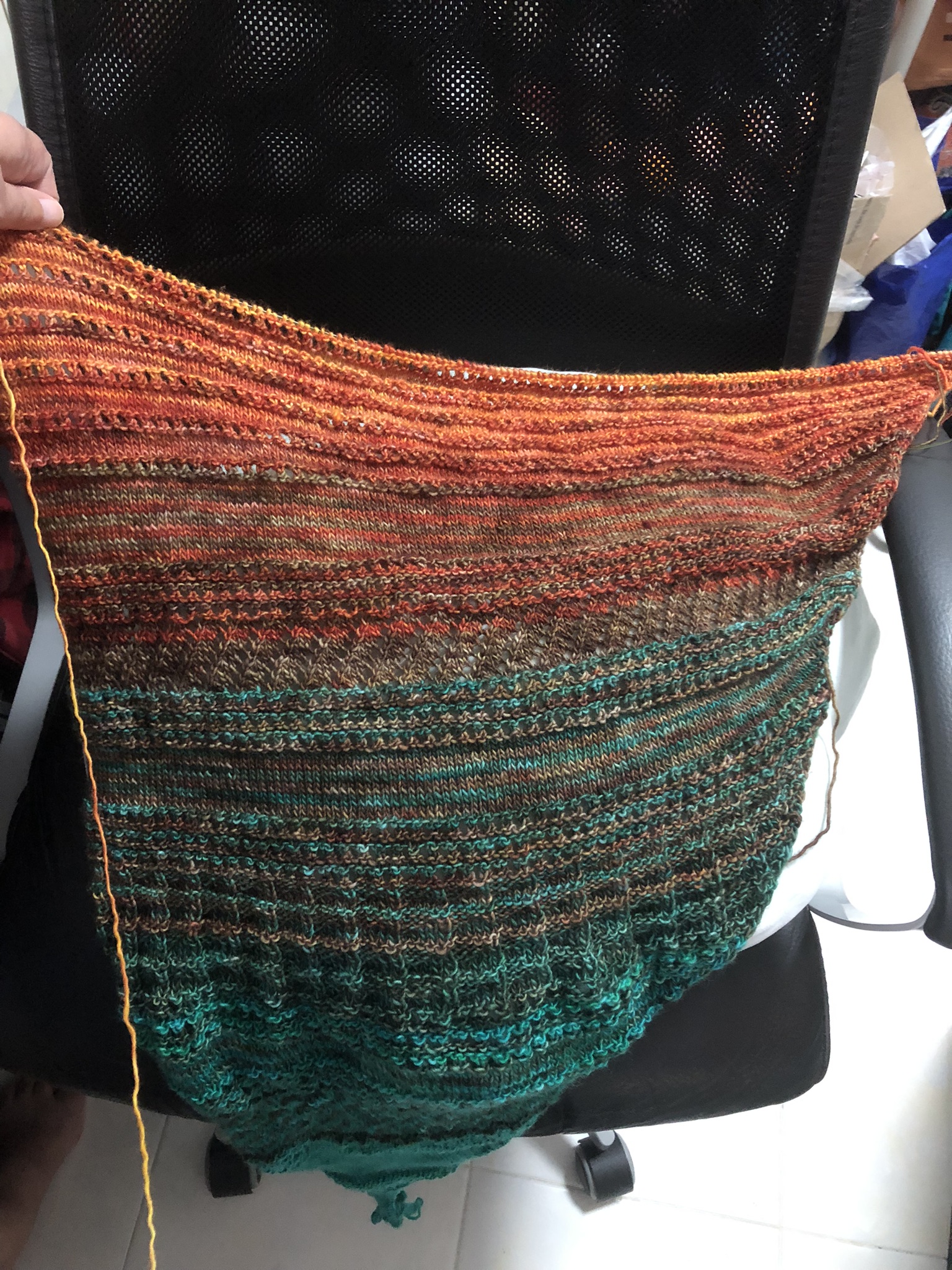Photo of a knitting project on the needles. It is triangular in shape, knitted in stripes in teal, greenish brown, reddish-brown, and red yarn. Orange yarn has been added in stripes in the newest sections, but it's not very obvious.