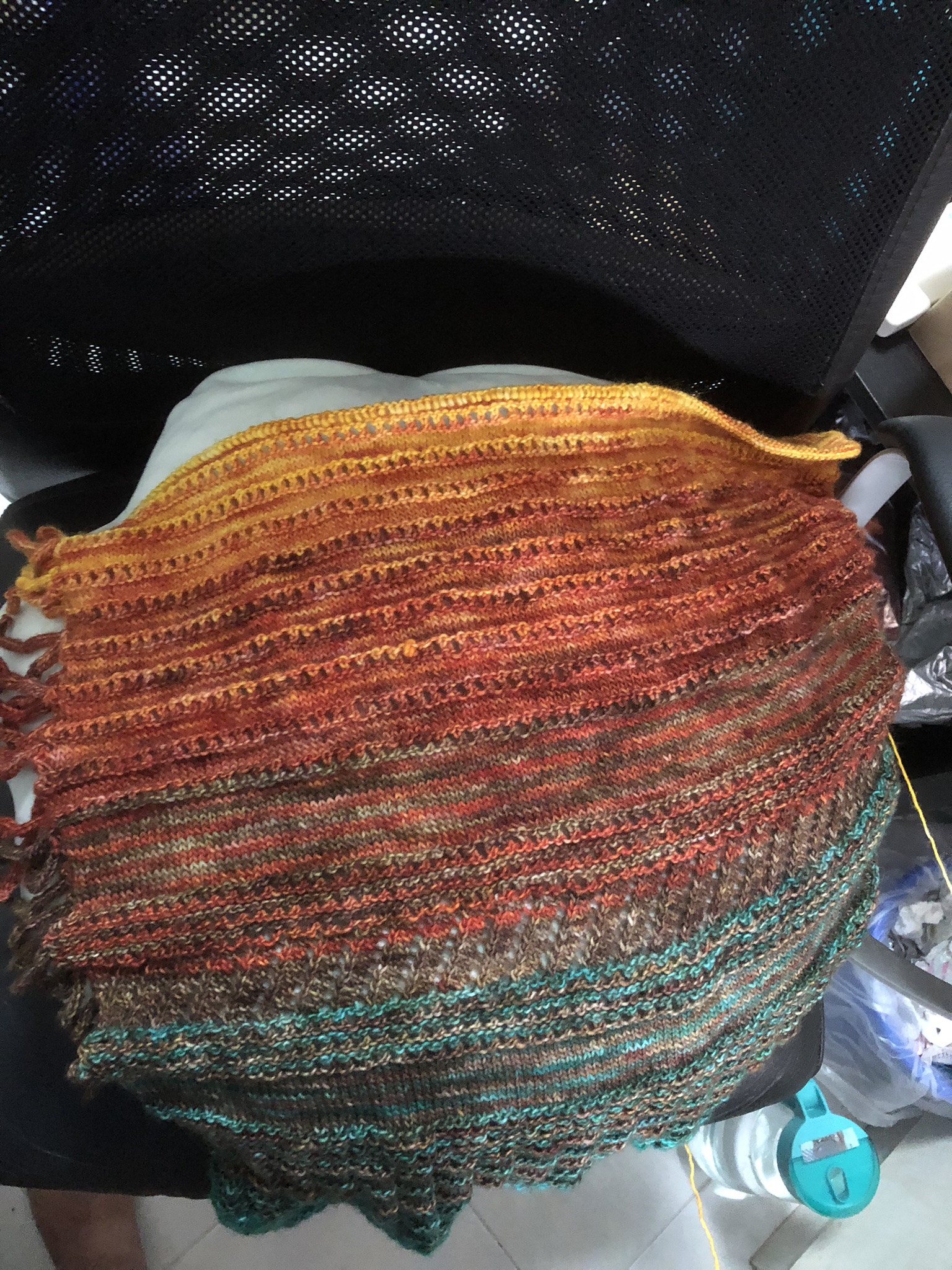 Photo of a knitting project on the needles. It is triangular in shape, knitted in stripes in teal, greenish brown, reddish-brown, red, and orange yarn. Yellow yarn has been added in stripes in the newest sections, but it's not very obvious.