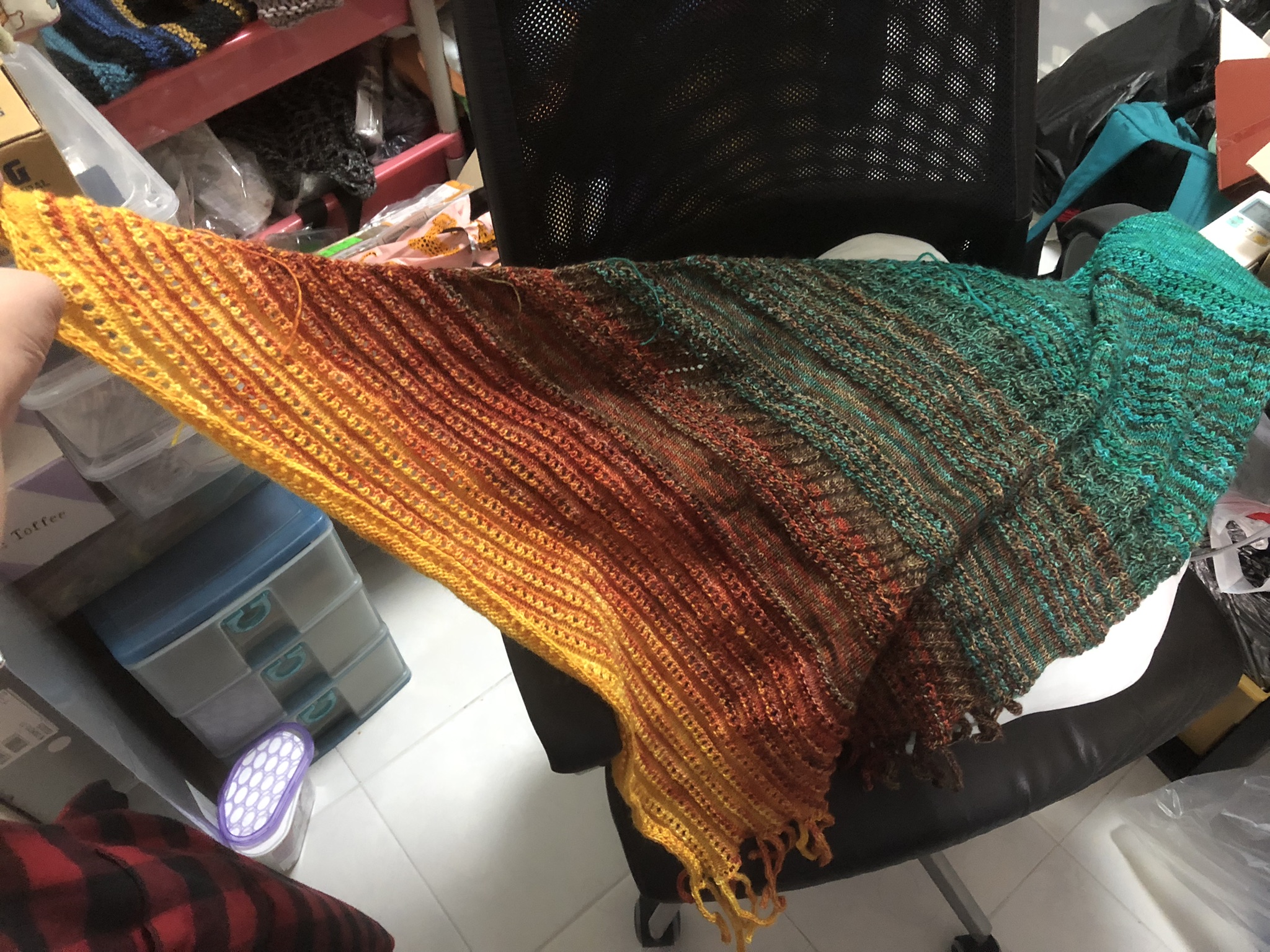 Photo of an almost complete knitting project. It is triangular in shape, knitted in stripes in teal, greenish brown, reddish-brown, red, orange, and yellow yarn. It is draped on a computer chair to show its shape.