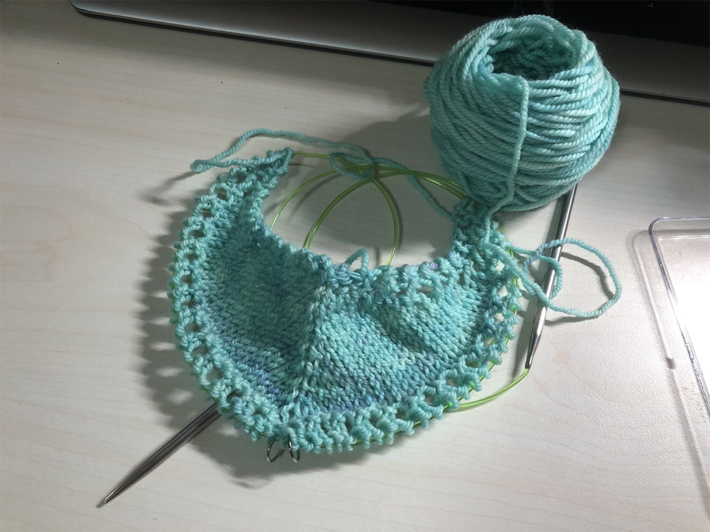 Photo of a knitting project on the needles. The yarn is a light cyan and the project is currently somewhat crescent-shaped.
