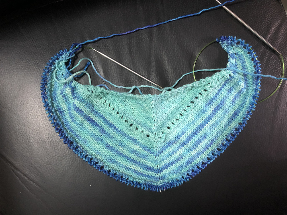 Photo of a knitting project on the needles. The project is currently somewhat crescent-shaped, like a triangle curling up at the ends. Most of the project is a light cyan, with some rows of darker blue striped in.