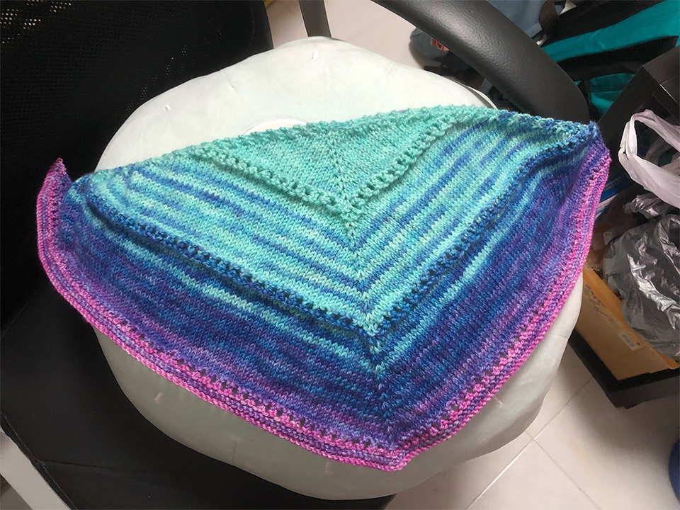 Photo of a knitting project. The project is a triangular kerchief, with the point of the triangle on the bottom, photographed spread out on a large round cushion placed on a black swivelling chair. Most of the project is a light cyan, with some rows of darker blue striped in, fading to purple and then pink on the outside edge.