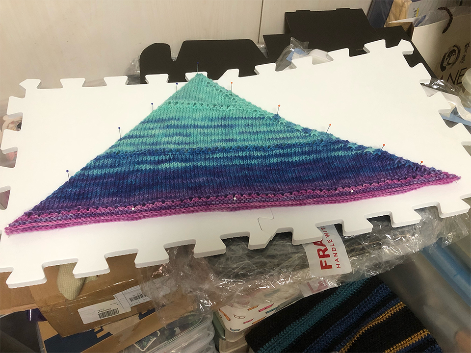 Photo of a knitting project. The project is a triangular kerchief, folded in half, pinned in place on two foam interlocking boards. Most of the project is a light cyan, with some rows of darker blue striped in, fading to purple and then pink on the bottom long edge. This setup is balanced on top of a stack of unassembled cardboard boxes and other storage boxes.
