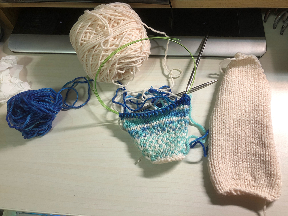 Photo of a knitting project on the needles on a desk. The piece currently being knitted is a small panel of spotted and striped blue and creamy white. There is a small blue ball of yarn and a larger ball of creamy white yarn above and left, and a larger vertically long panel of knitted creamy white fabric to the right.