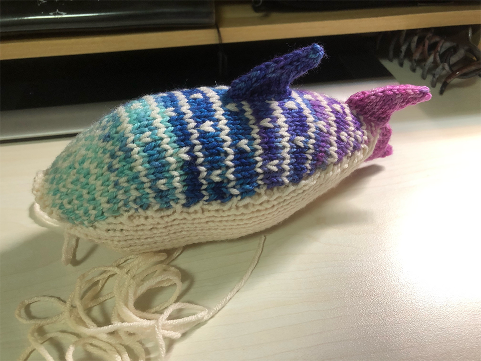 Photo of an incomplete knitted whale shark plush on a desk facing the left. It is light cyan to blue to purple to pink starting from the head to the tail, with a creamy white underside and stripes and spots. It has a dorsal fin and the tail fins, is stuffed, but doesn't have the front fins and eyes, and the front of the face is still open, with a long thread of creamy white yarn attached.