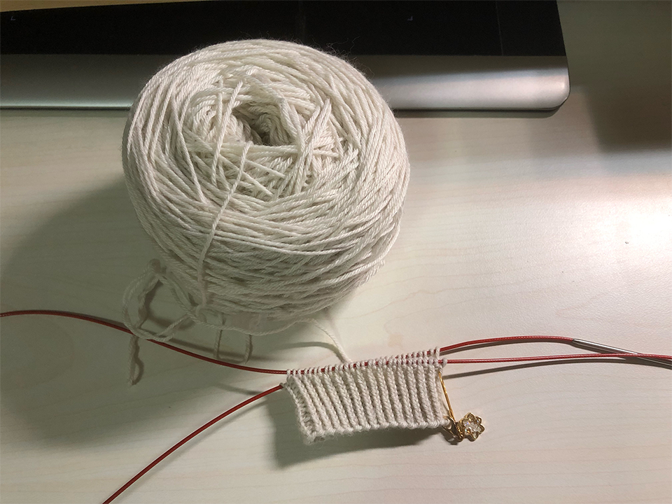 Photo of a knitting project on the needles on a desk. There is a large ball of greyish-white yarn placed above it. The knitted portion is a part of a ribbed cuff in neat vertical rows. A small konpeito-shaped enamel charm is attached to the bottom right corner.