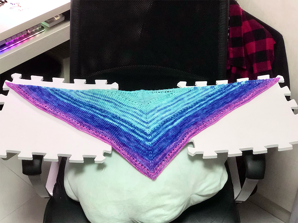Photo of a triangular kerchief knitted in a gradient of cyan to blue to purple to pink, spread on foam puzzle panels and a large cushion balanced on an office chair.