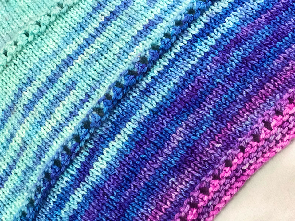 Closeup photo of knitted fabric against a creamy white backdrop. The fabric is in a striped gradient from cyan to blue to purple to pink, with a few rows of eyelets.