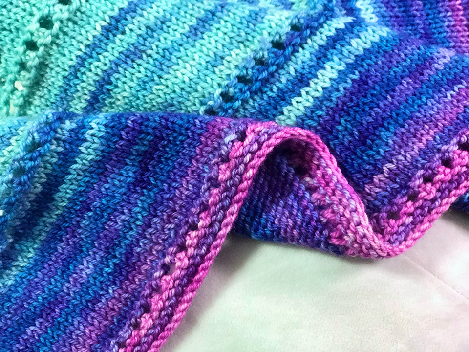 Closeup photo of knitted fabric against a creamy white backdrop, folded a few times. The fabric is in a striped gradient from cyan to blue to purple to pink, with a few rows of eyelets.