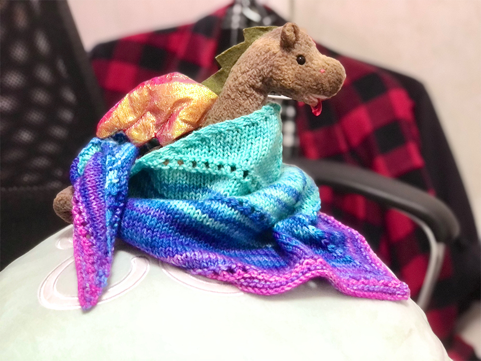 Photo of a triangular kerchief wrapped around a beanie baby Scorch. The kerchief is in a striped gradient of cyan to blue to purple to pink and tied around the back. The entire setup is photographed on a large moon jelly cushion on an office chair.