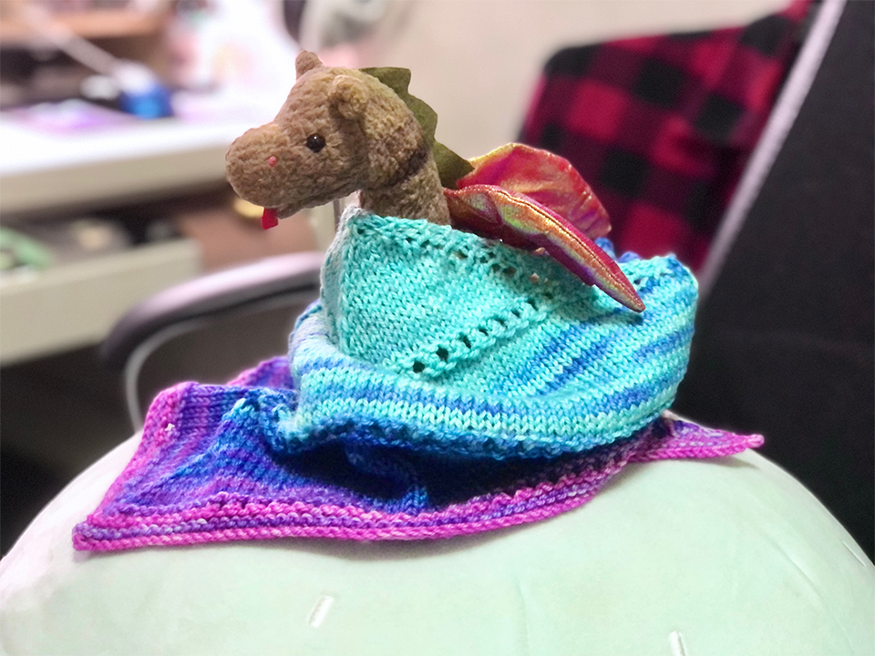 Photo of a triangular kerchief wrapped around a beanie baby Scorch. The kerchief is in a striped gradient of cyan to blue to purple to pink and tied around the back. The entire setup is photographed on a large moon jelly cushion on an office chair.