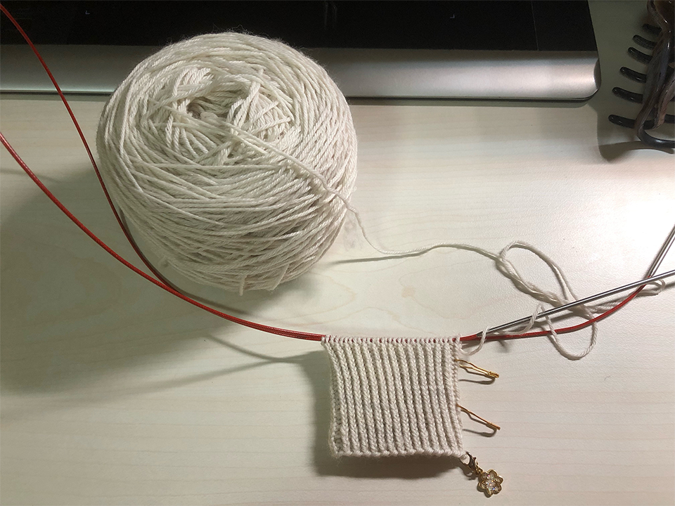 Photo of a knitting project on the needles on a desk. There is a large ball of greyish-white yarn placed above it. The knitted portion is a part of a ribbed cuff in neat vertical rows. A small konpeito-shaped enamel charm is attached to the bottom right corner.