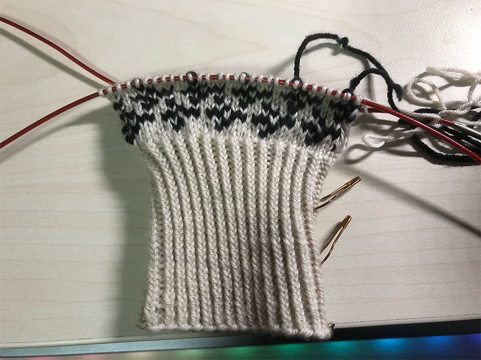 Photo of a knitting project on the needles on a desk. The knitted portion is a part of a ribbed cuff in neat vertical rows, with two rows of bird footprints above the cuff.
