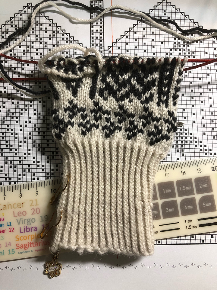 Photo of a knitting project on the needles on a desk. The knitted portion shows a ribbed cuff in neat vertical rows, with two rows of bird footprints above the cuff and an emerging pattern above that. Underneath the project is a white ruler with a design cheatsheet printed on it and the printed knitting chart itself.