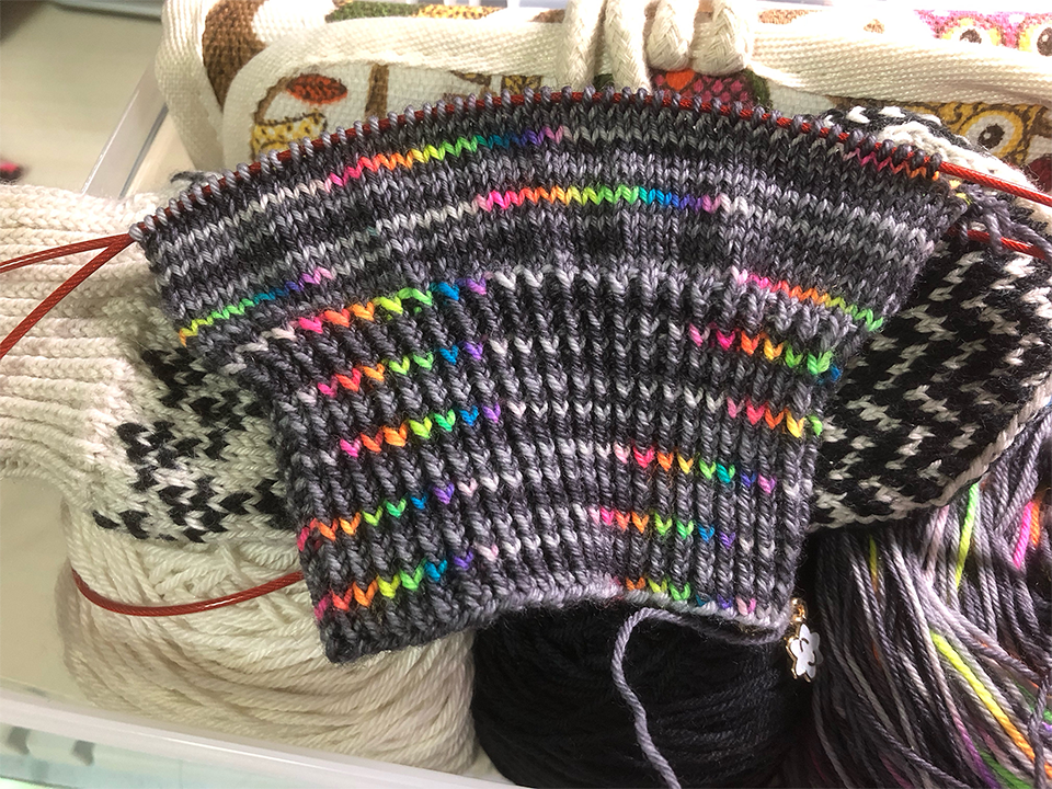 Photo of a knitting project on the needles. The project is a sock in greys and rainbow yarn. The cuff has been knitted along with a few rows of the leg. This is placed on top of a box containing several balls of yarn.