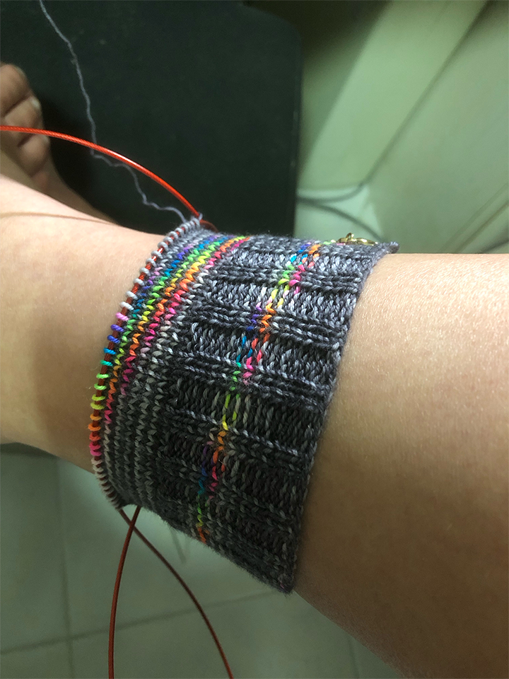 Photo of a knitting project on the needles. The project is a sock in greys and rainbow yarn. The cuff has been knitted along with a few rows of the leg. It has been put on a leg to test the fit.
