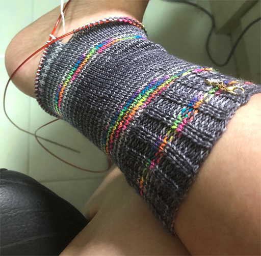 Photo of a knitting project on the needles. The project is a sock in greys and rainbow yarn. The cuff and leg has been knitted, along with a few rows of the heel. It has been put on a leg to test the fit.