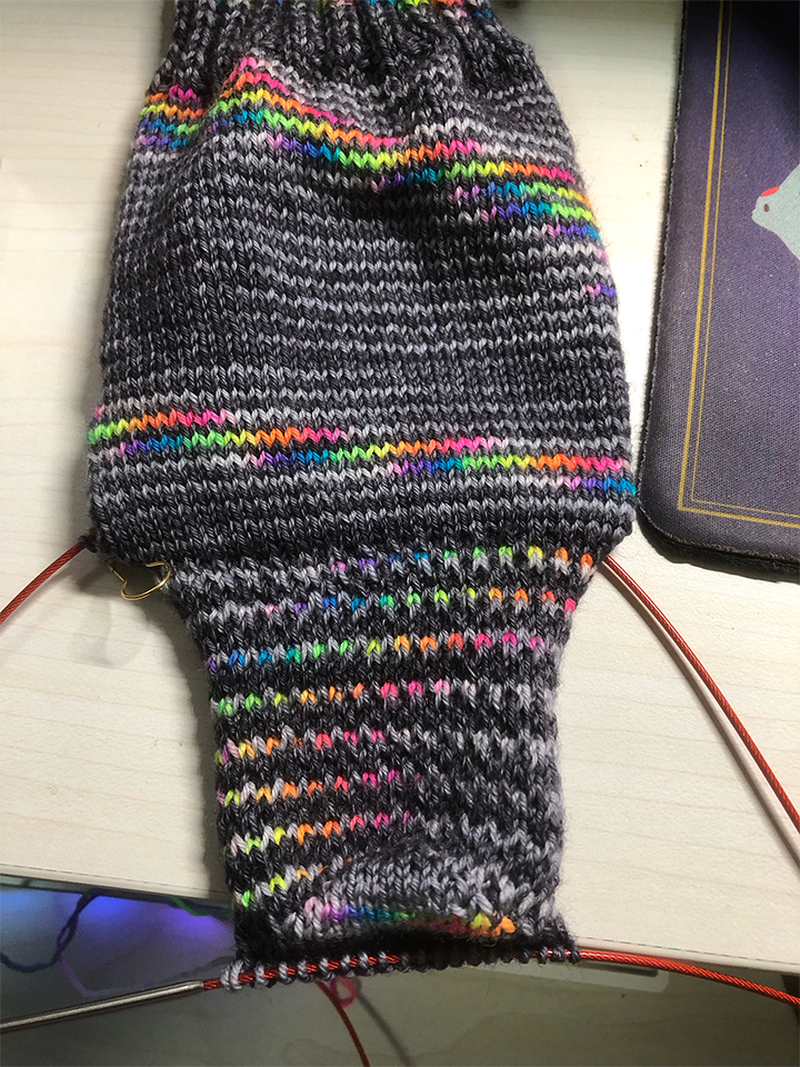 Photo of a knitting project on the needles. The project is a sock in greys and rainbow yarn. The cuff and leg has been knitted, along with the rest of the heel. It is placed flat on a table to show the heel.